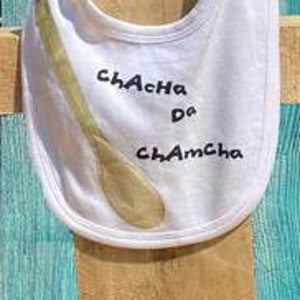 以下が含まれることがあります： 「ChaCha da ChamCha」の黒い筆記体と木のスプーンのグラフィックが特徴の白いベビービブ。ビブは柔らかい生地で作られており、曲線的なネックラインとスナップ留めが付いています。