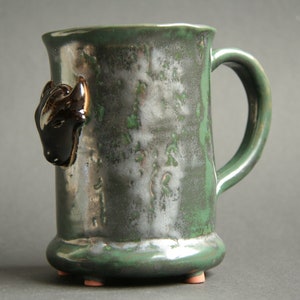 May include: A green ceramic mug with a black and white animal head on the side. The mug has a handle and a rounded base with three small feet.