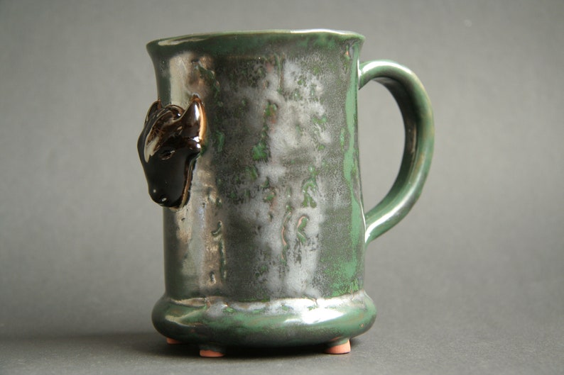 May include: A green ceramic mug with a black and white animal head on the side. The mug has a handle and a rounded base with three small feet.