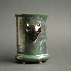 May include: A green ceramic vase with a black and white buffalo head design. The vase has a rounded bottom and three small feet.