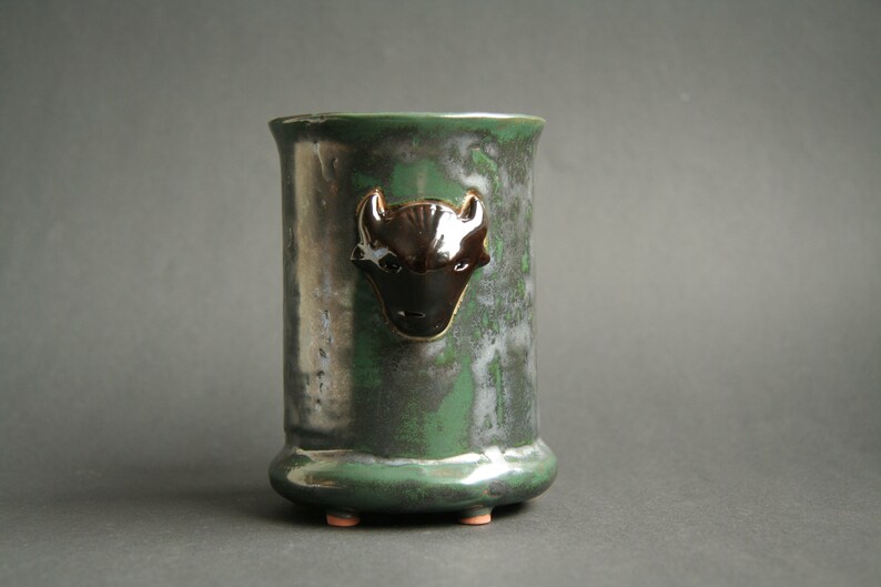 May include: A green ceramic vase with a black and white buffalo head design. The vase has a rounded bottom and three small feet.