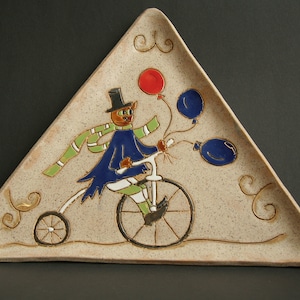 Pode incluir: Uma travessa de cerâmica triangular com um design extravagante. Apresenta um gato de cartola andando de bicicleta e segurando balões. O gato usa um casaco azul e um cachecol verde. A travessa tem um fundo bege salpicado.
