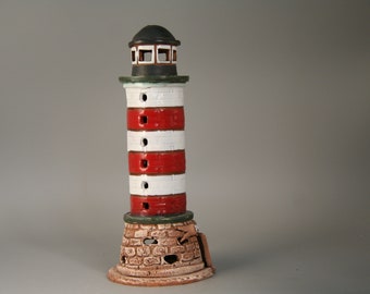 May include: A ceramic lighthouse figurine with red and white stripes, a green base, and a black roof. The base is textured to look like stone.