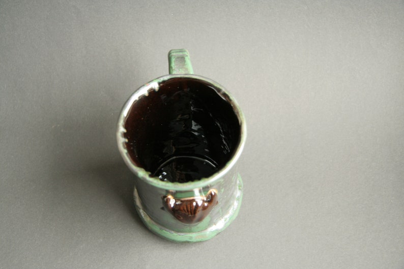 May include: A green ceramic mug with a brown interior and a brown animal figurine on the front.
