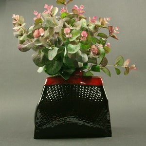May include: A black and red ceramic vase with a textured surface, filled with a bouquet of green leaves and pink flowers.