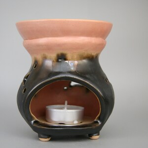 以下が含まれることがあります： ティーライトキャンドル用の小さな丸い開口部が付いた、黒とピンクのセラミック製オイルバーナー。バーナーは、カットアウトとテクスチャ加工された仕上げで装飾的なデザインになっています。