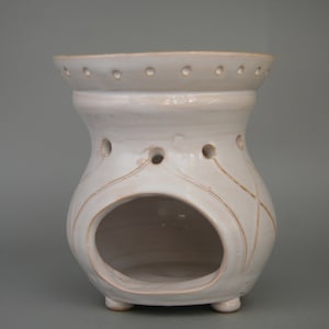 Può includere: Bruciatore per olio in ceramica bianca con un design decorativo e una grande apertura al centro. Il bruciatore ha un bordo rialzato con piccoli fori attorno alla parte superiore.