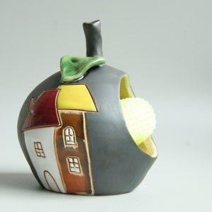 May include: A gray ceramic apple-shaped dish with a house painted on the side. The dish has a cutout in the side to hold a sponge or scrubber. The house is painted in shades of red, yellow, brown, and white.