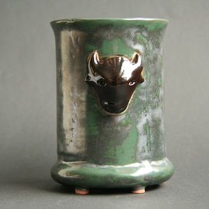 May include: A green ceramic vase with a black and white animal head design. The vase has a rounded bottom and three small feet.