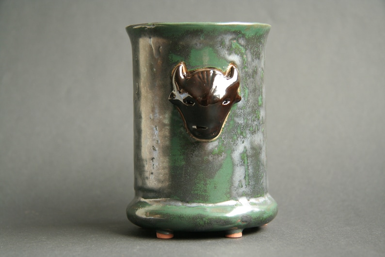 May include: A green ceramic vase with a black and white animal head design. The vase has a rounded bottom and three small feet.