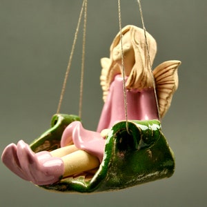 Puede incluir: Una figurita de ángel de cerámica con cabello rubio y alas rosadas, sentada en un columpio verde. El ángel lleva un vestido rosa y tiene una dulce expresión en su rostro.