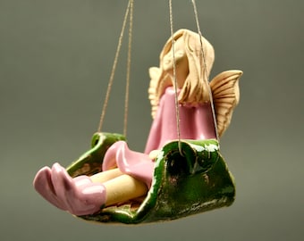May include: A ceramic angel figurine with blonde hair and pink wings, sitting on a green swing. The angel is wearing a pink dress and has a sweet expression on her face.