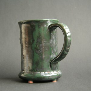 May include: A green ceramic mug with a textured glaze and a handle. The mug has a rounded base with three small feet.