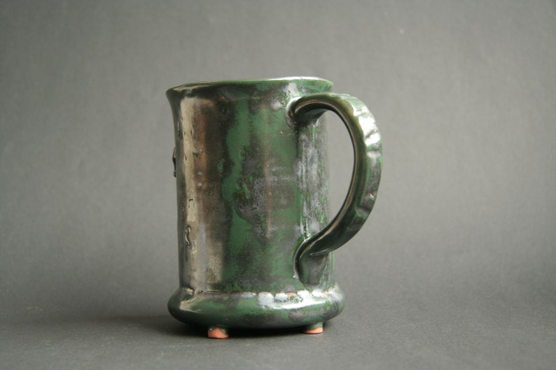 May include: A green ceramic mug with a textured glaze and a handle. The mug has a rounded base with three small feet.