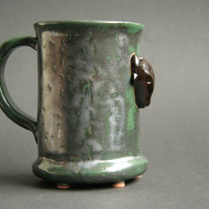 May include: A green ceramic mug with a black animal head on the side. The mug has a textured surface and a handle.