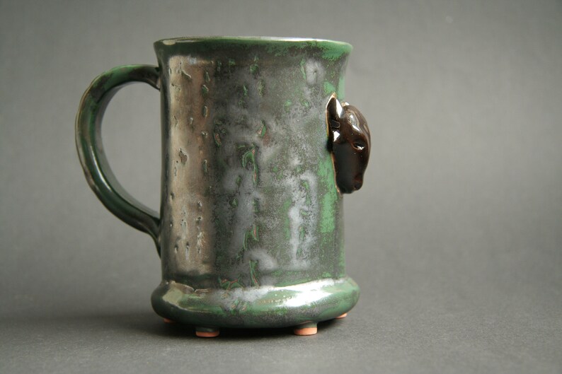 May include: A green ceramic mug with a black animal head on the side. The mug has a textured surface and a handle.