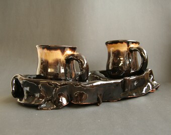 May include: Two brown and white ceramic mugs with black glaze sit on a black ceramic tray with a wavy, organic shape.