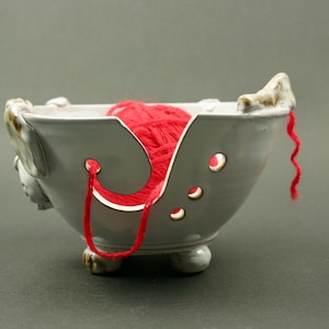 May include: White ceramic yarn bowl with a red ball of yarn inside. The bowl has holes for the yarn to pass through and a decorative design on the side.