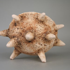 Puede incluir: Una escultura de cer&aacute;mica de un objeto redondo y espinoso con una superficie rugosa y texturizada. El objeto es de color marr&oacute;n claro con acentos blancos.