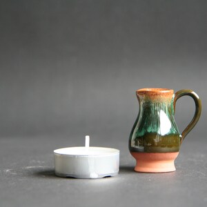 Pode incluir: Um pequeno jarro de cerâmica marrom e verde com alça, ao lado de uma vela de chá branca em um suporte prateado.
