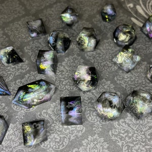 May include: A collection of iridescent resin dice in various geometric shapes, including cubes, pyramids, and multi-sided dice. The dice have a black and white marbled effect with hints of blue, green, and yellow. Some dice have visible numbers.
