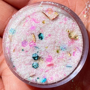 May include: A small, clear container filled with a mix of pink, white, and iridescent glitter. The glitter is interspersed with colourful, geometric shapes and small, gold-coloured flakes. The container is held in a hand.