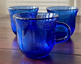 May include: Three cobalt blue glass cups with a textured design. The cups have a rounded shape and a handle.