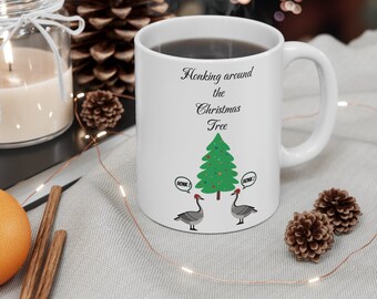 May include: White ceramic coffee mug with a Christmas tree and two geese with red Santa hats. The text on the mug reads "Honking around the Christmas Tree".