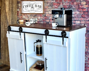 Puede incluir: Armario de cocina blanco con puertas correderas de granero, un letrero de bar de café, una cafetera y una jarra de café. El armario tiene dos estantes y una encimera de madera.