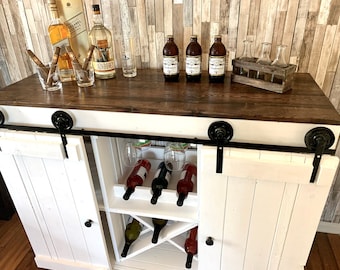 May include: White bar cabinet with sliding barn doors, a brown countertop, and a wine rack. The cabinet has black hardware and is filled with wine bottles and glasses.