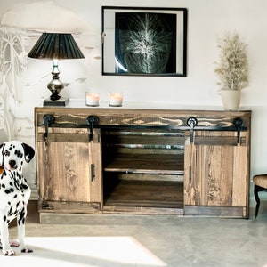 May include: A rustic wooden cabinet with sliding doors and black hardware. A Dalmatian dog sits in front of the cabinet. The cabinet has a lamp, candles, and a framed picture on top. A decorative chair is to the right.
