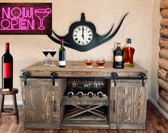May include: A rustic wooden bar cabinet with sliding doors, a wine rack, and glass holders. The cabinet has a dark brown finish and features black metal hardware. There are two bottles of liquor, two glasses of whiskey, and a bottle of wine on the counter.