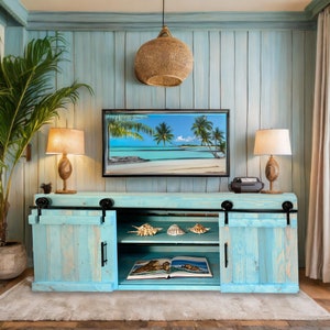 May include: A light blue wooden TV stand with sliding barn doors. The stand has two shelves and is decorated with seashells. The stand is in front of a light blue wall with a tropical beach scene on the TV.