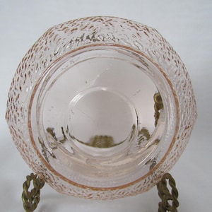 May include: A vintage pink glass bowl with a textured exterior and a smooth interior. The bowl is supported by a decorative metal stand. The glass has a delicate, translucent quality, and the stand has a twisted rope design.