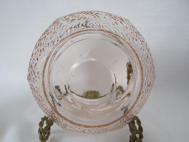 May include: A vintage pink glass bowl with a textured exterior and a smooth interior. The bowl is supported by a decorative metal stand. The glass has a delicate, translucent quality, and the stand has a twisted rope design.