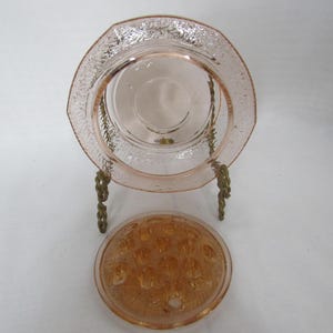 May include: A pink glass plate with a decorative floral pattern and a matching flower frog. The plate is displayed on a gold-colored metal stand. The flower frog is round with multiple holes.