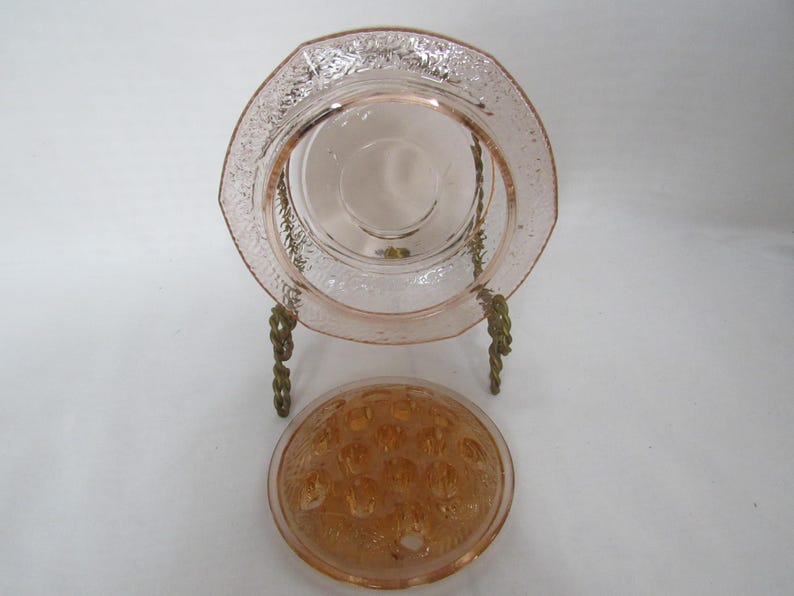 May include: A pink glass plate with a decorative floral pattern and a matching flower frog. The plate is displayed on a gold-colored metal stand. The flower frog is round with multiple holes.