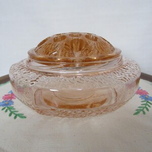 May include: A vintage peach-colored glass flower frog with a textured rim and a domed lid featuring multiple holes. The flower frog sits on a white embroidered cloth with floral designs.