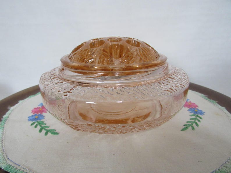 May include: A vintage peach-colored glass flower frog with a textured rim and a domed lid featuring multiple holes. The flower frog sits on a white embroidered cloth with floral designs.