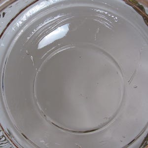 May include: A clear glass bowl with a decorative, twisted gold-colored rim. The bowl has a circular design with a central indentation. The glass is transparent, allowing light to pass through, and the rim has a textured appearance.