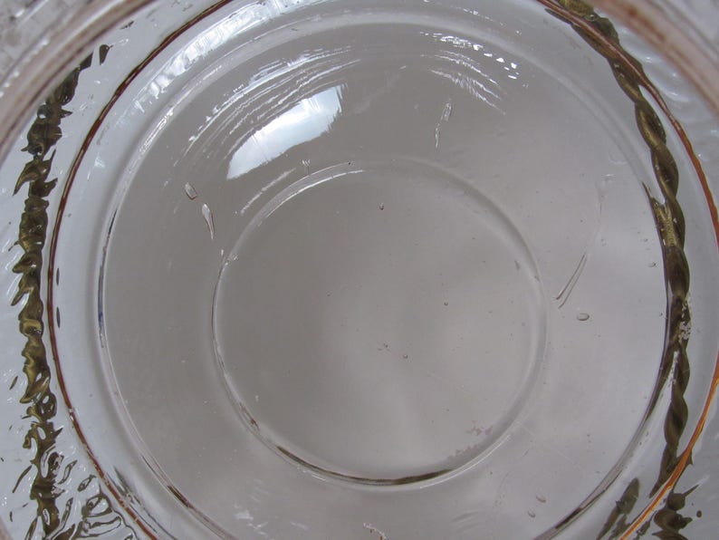 May include: A clear glass bowl with a decorative, twisted gold-colored rim. The bowl has a circular design with a central indentation. The glass is transparent, allowing light to pass through, and the rim has a textured appearance.
