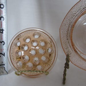 May include: A light pink glass flower frog with multiple holes, next to a matching dish. The flower frog is approximately 3 inches in diameter. The dish has a decorative rim. Both items are displayed on a white surface.