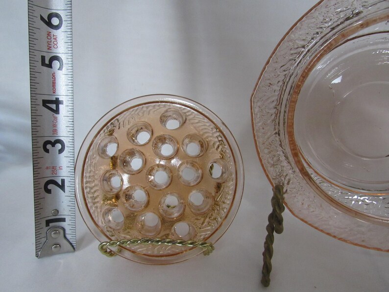 May include: A light pink glass flower frog with multiple holes, next to a matching dish. The flower frog is approximately 3 inches in diameter. The dish has a decorative rim. Both items are displayed on a white surface.
