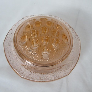 May include: A vintage, light pink glass flower frog with a round, textured base and a domed top. The top features multiple holes for arranging flowers. The edges of the base have a decorative, etched pattern.