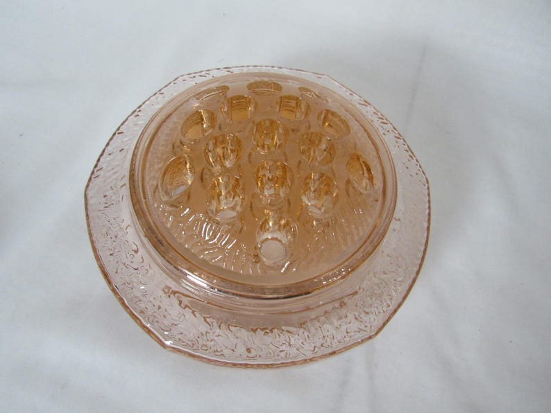 May include: A vintage, light pink glass flower frog with a round, textured base and a domed top. The top features multiple holes for arranging flowers. The edges of the base have a decorative, etched pattern.