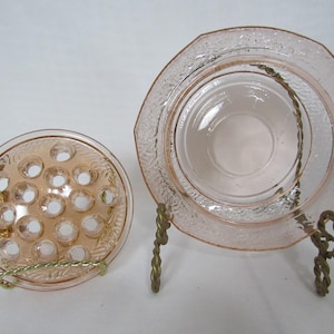 May include: Two vintage pink glass items: a flower frog and a small plate with a textured rim, displayed on decorative metal stands. The flower frog has multiple holes for arranging flowers.