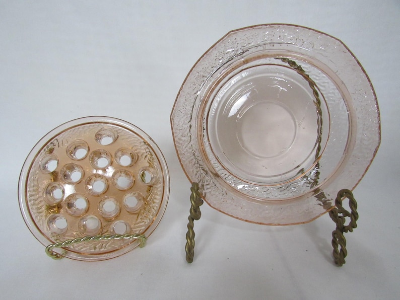 May include: Two vintage pink glass items: a flower frog and a small plate with a textured rim, displayed on decorative metal stands. The flower frog has multiple holes for arranging flowers.