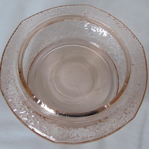 May include: A vintage pink glass bowl with a textured, octagonal rim. The bowl has a circular interior and a slightly raised base. The glass has a subtle, translucent quality, suggesting it is likely a decorative or serving piece.
