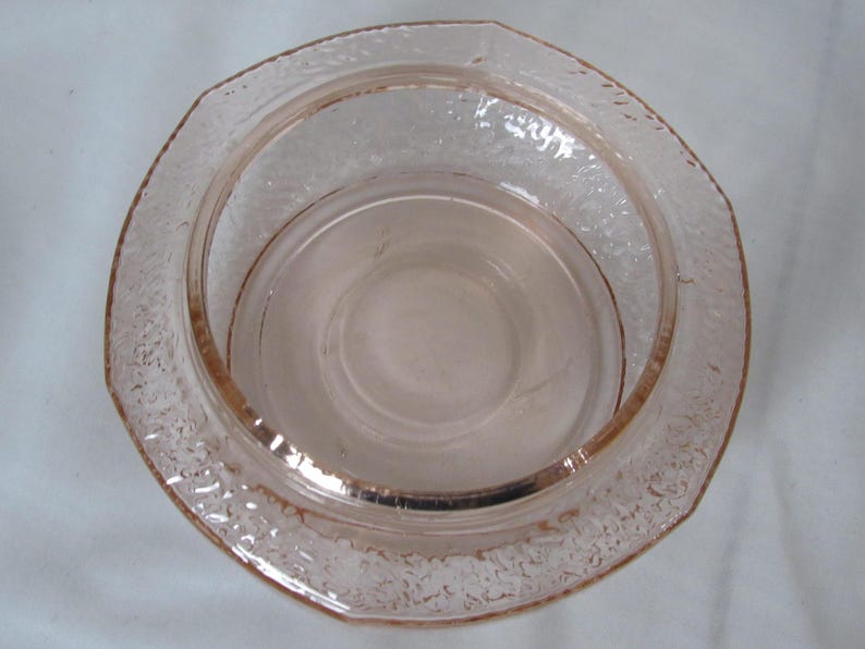 May include: A vintage pink glass bowl with a textured, octagonal rim. The bowl has a circular interior and a slightly raised base. The glass has a subtle, translucent quality, suggesting it is likely a decorative or serving piece.
