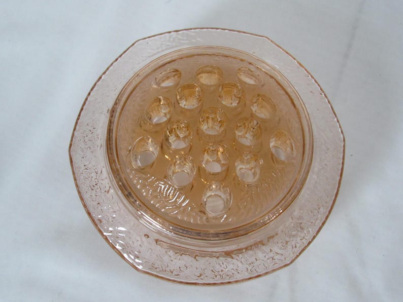 May include: A vintage, light pink glass flower frog with a textured, octagonal rim. The center features multiple holes for arranging flowers. The glass has a subtle, translucent quality, and the overall design is simple and elegant.
